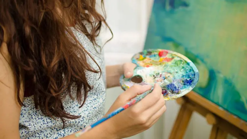 Person with long hair holding a paint palette and brush, painting on a canvas on an easel.