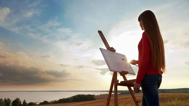 A person with long hair stands outdoors at an easel, painting on a canvas, with a view of the horizon, clouds, and fields in the background.